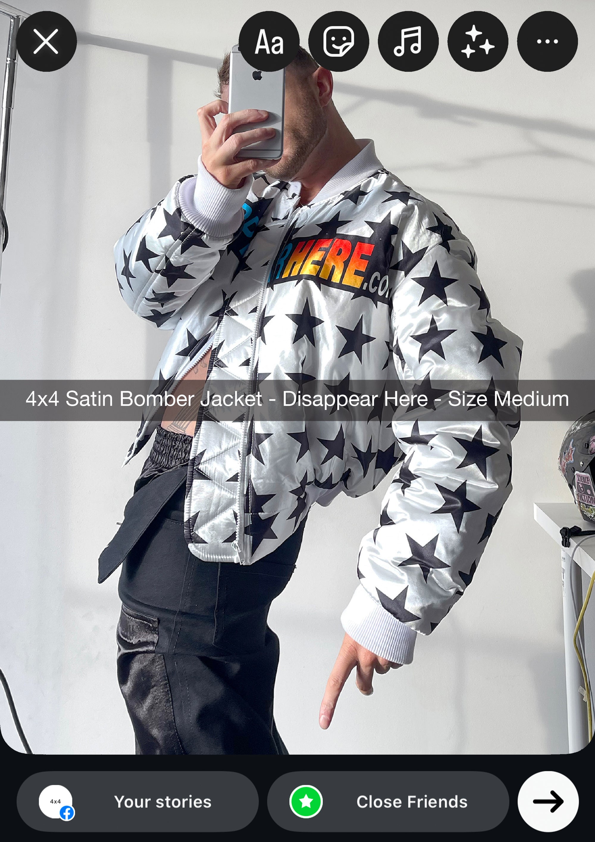Person wearing a satin bomber jacket with star pattern and colorful text, taking a mirror selfie.