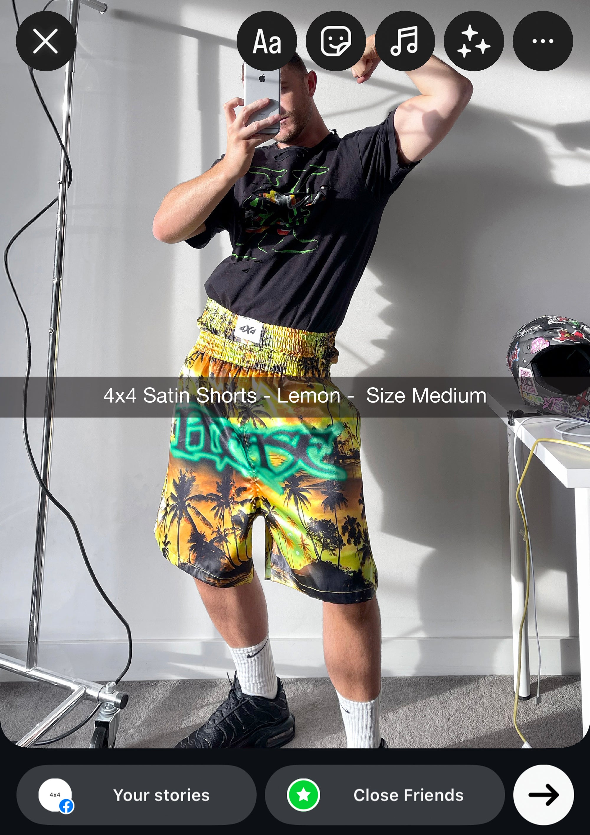 Person wearing colorful shorts taking a mirror selfie in a room with a white wall and gray floor.